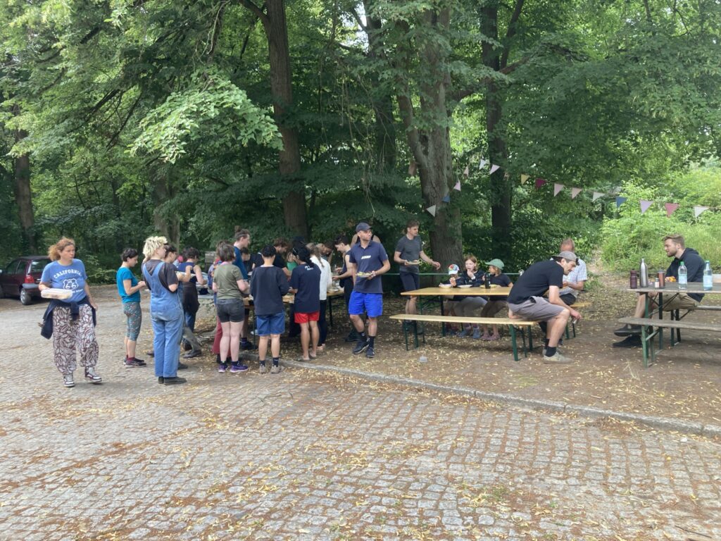 Unter schattigen Bäumen und Wimpelkette stärken sich die Helfer:innen der Solawi Spörgelhof am Mitbringbuffet – Gemeinschaft, die satt macht.